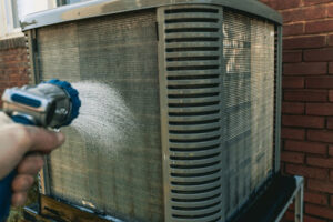 A hand using a hose to clean an outdoor AC unit.