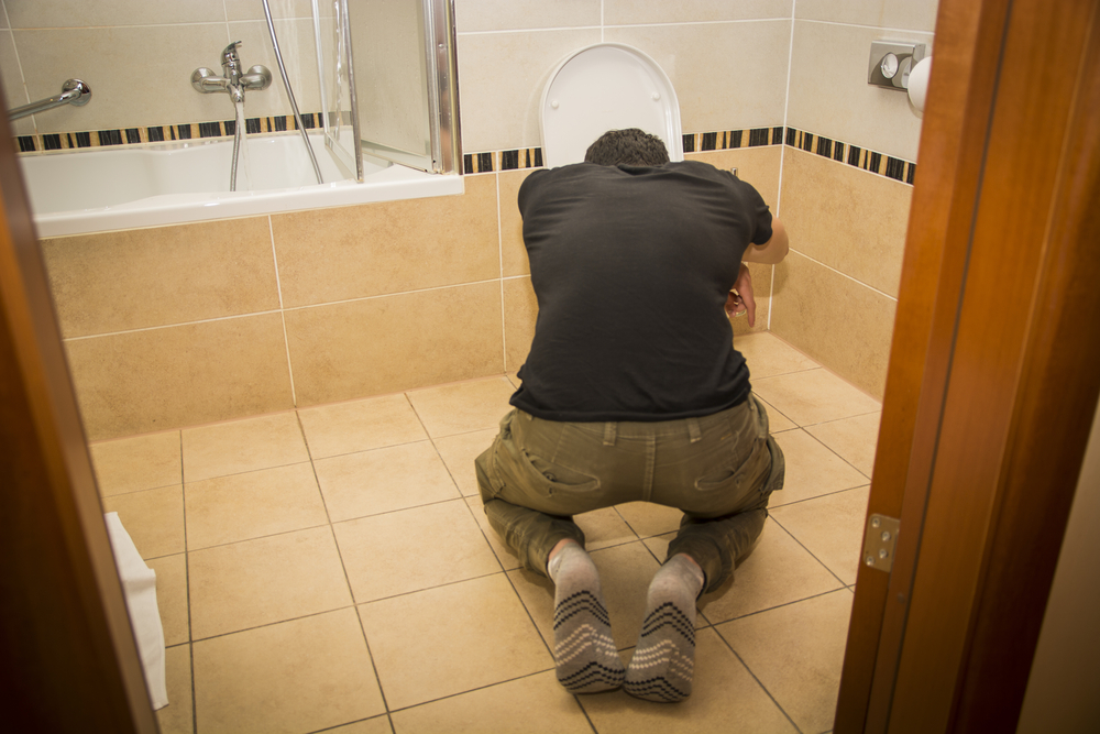 Rear View of a Drunk Young Man Vomiting in the Toilet at Home While in Kneeling Position.